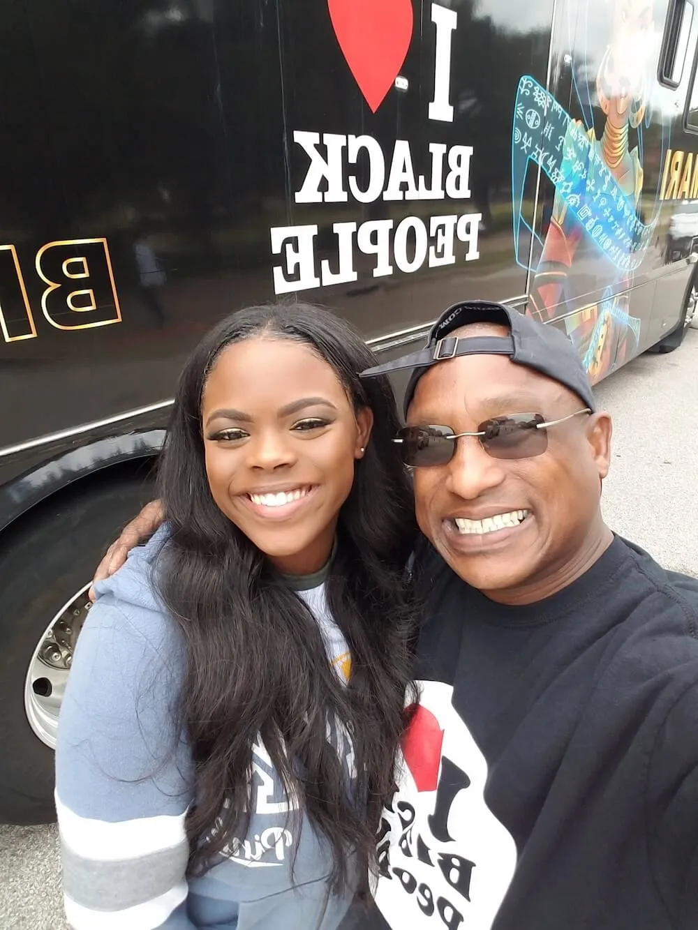 Smiling man and woman taking a selfie in front of a black vehicle with 'I ❤️ Black People' written on it.