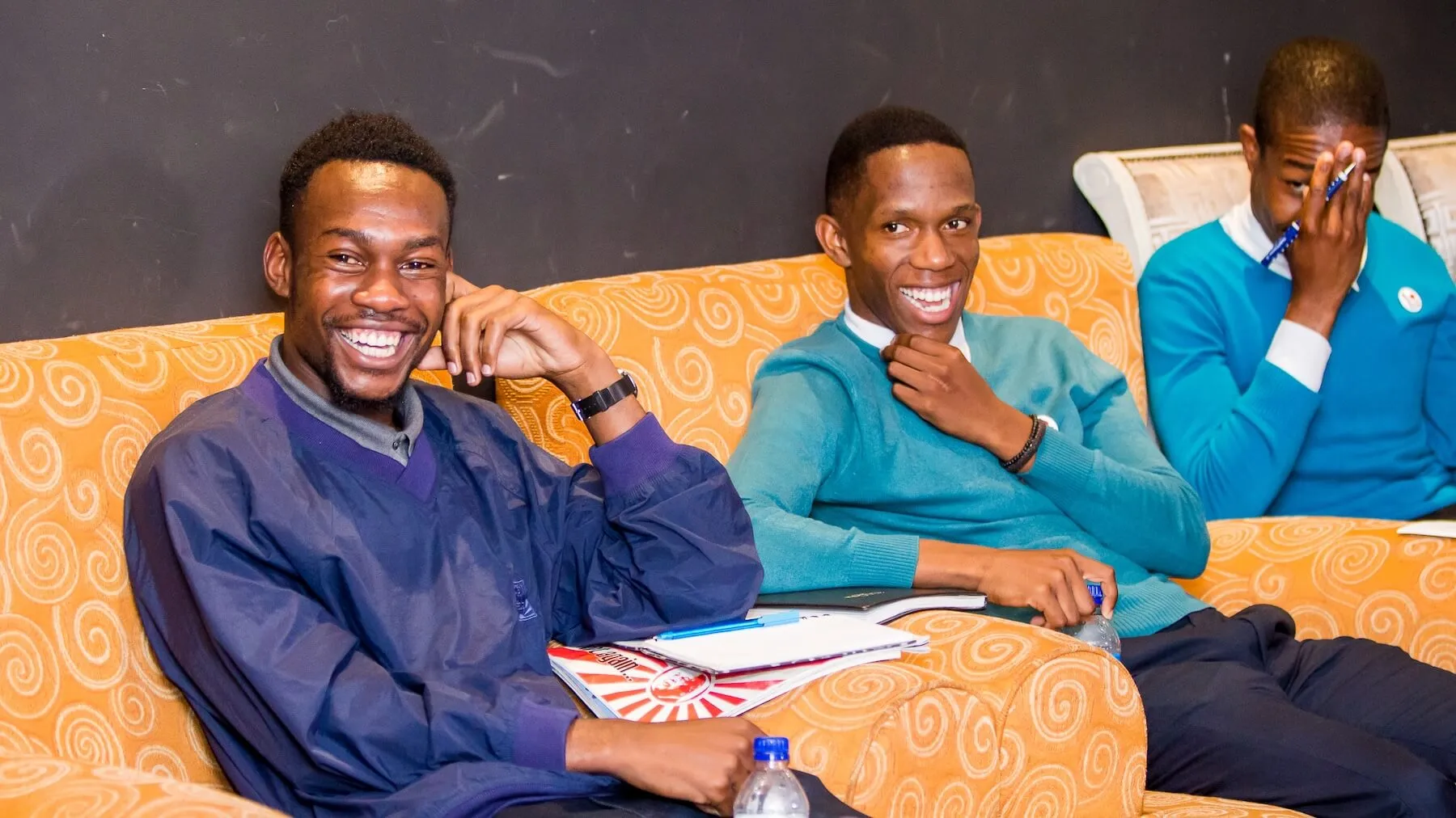 Three young men sitting on orange patterned couches, smiling and laughing, with notebooks and water bottles.