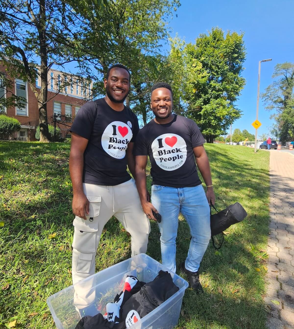 Two smiling men wearing black T-shirts that say 'I love Black People' standing outdoors on grass next to a clear plastic bin with similar T-shirts inside.