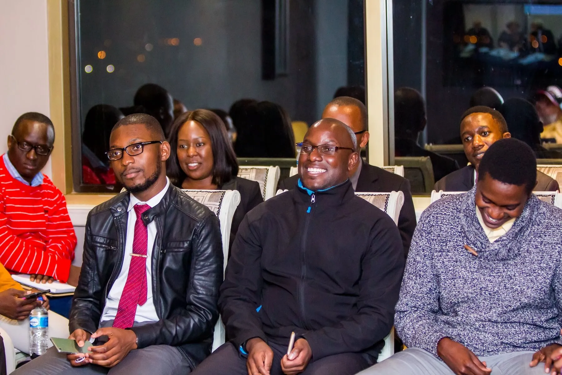 A group of people seated indoors, some smiling and engaged, including a man in a black leather jacket and red tie, and another man in a black jacket holding a phone.