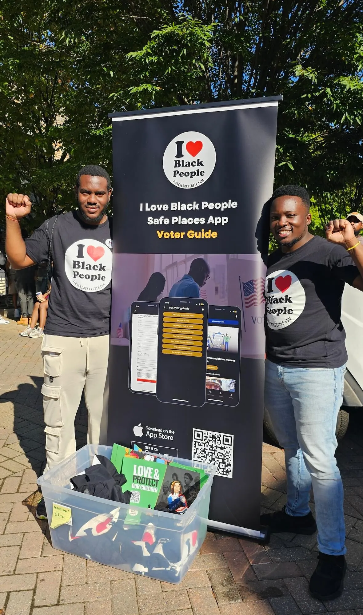 Two men wearing black 'I Love Black People' t-shirts raising fists beside a banner promoting the 'I Love Black People Safe Places App Voter Guide' with app download QR code and a plastic bin of merchandise.