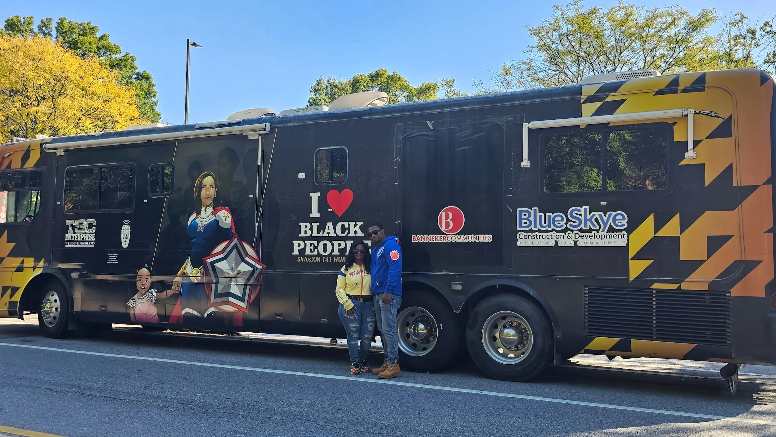 Two people standing in front of a large black and yellow bus with logos including 'I love Black People,' 'Banneker Communities,' and 'Blue Skye Construction & Development.'