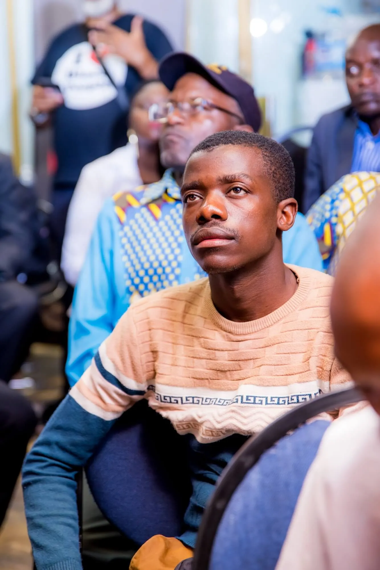 Young man in a patterned sweater attentively listening at an indoor event with others seated behind him.