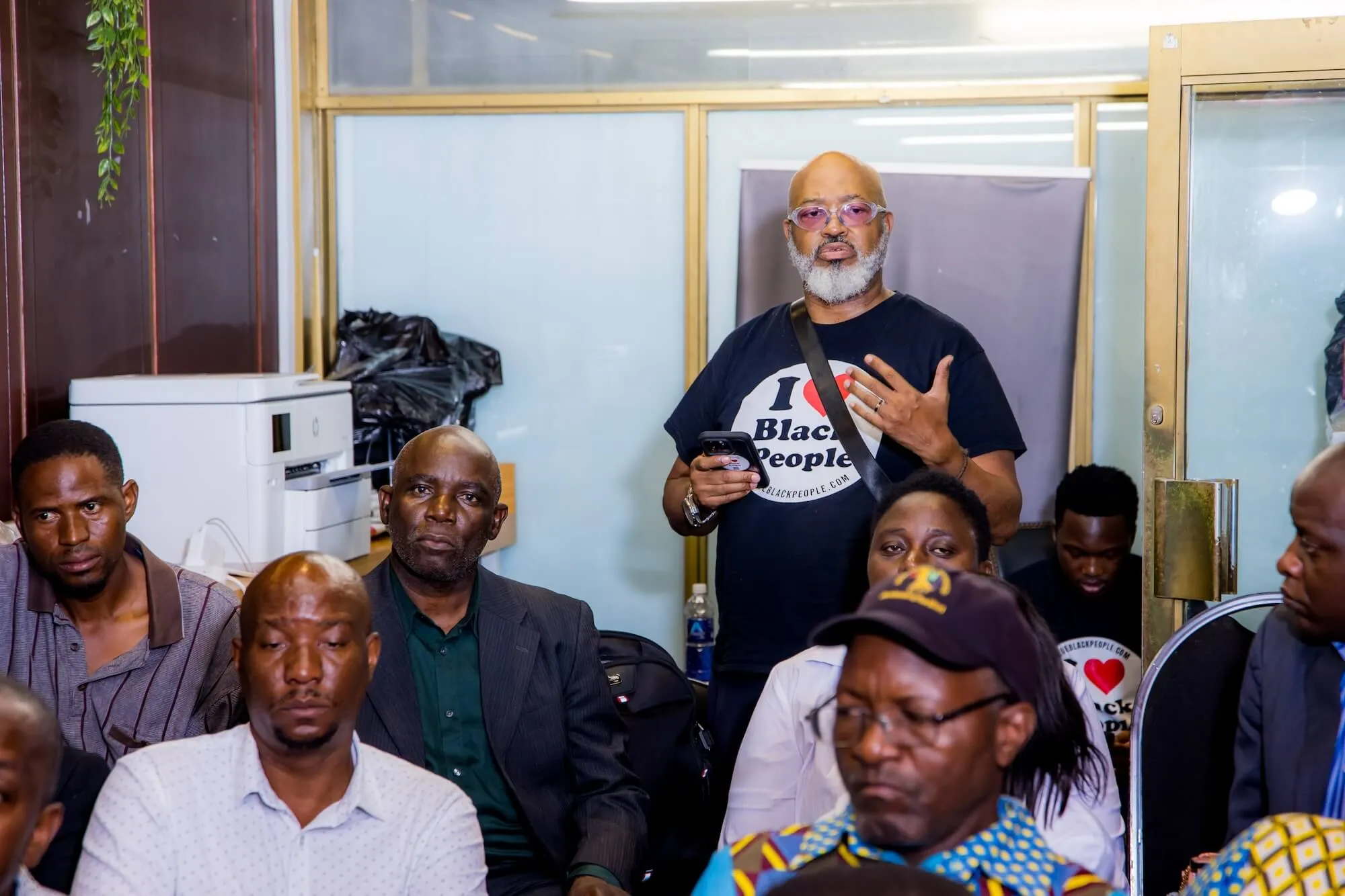 A man wearing sunglasses and an 'I love Black People' T-shirt stands speaking in a room filled with seated attentive people.