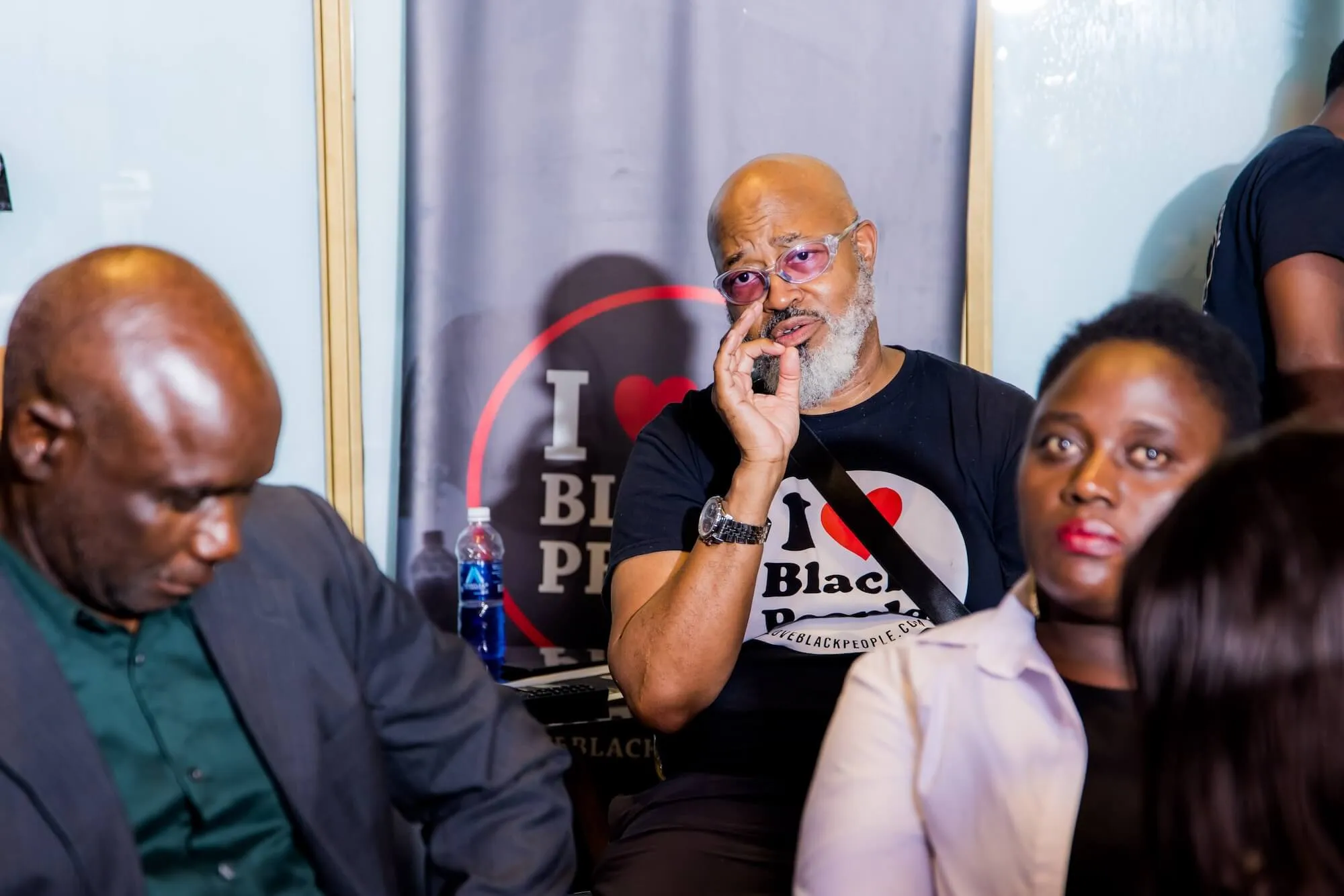 Man with gray beard and purple glasses wearing an 'I love Black People' t-shirt, seated among others at an indoor event.