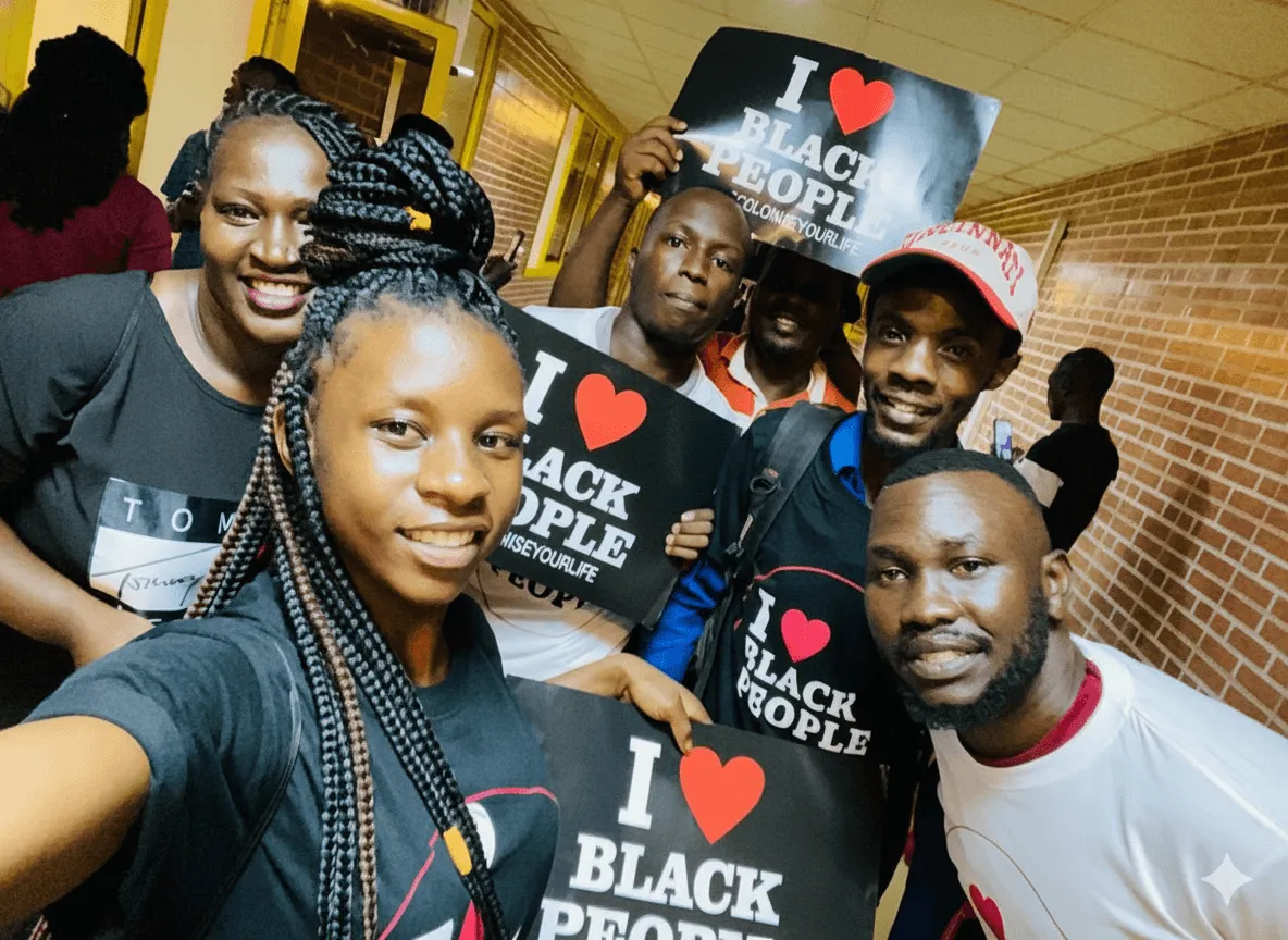 Group of smiling Black people holding signs that say 'I ♥ Black People' and wearing matching T-shirts in a hallway with brick walls.