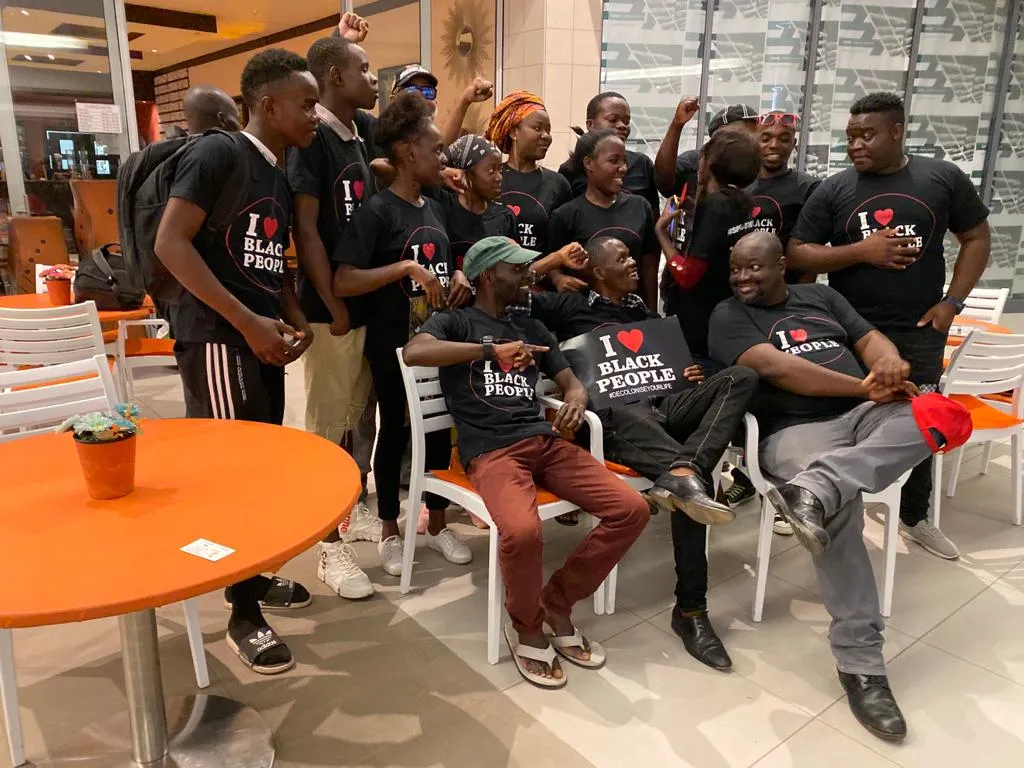 Group of people wearing matching black T-shirts with 'I ❤️ Black People' text, posing cheerfully indoors near orange tables and white chairs.