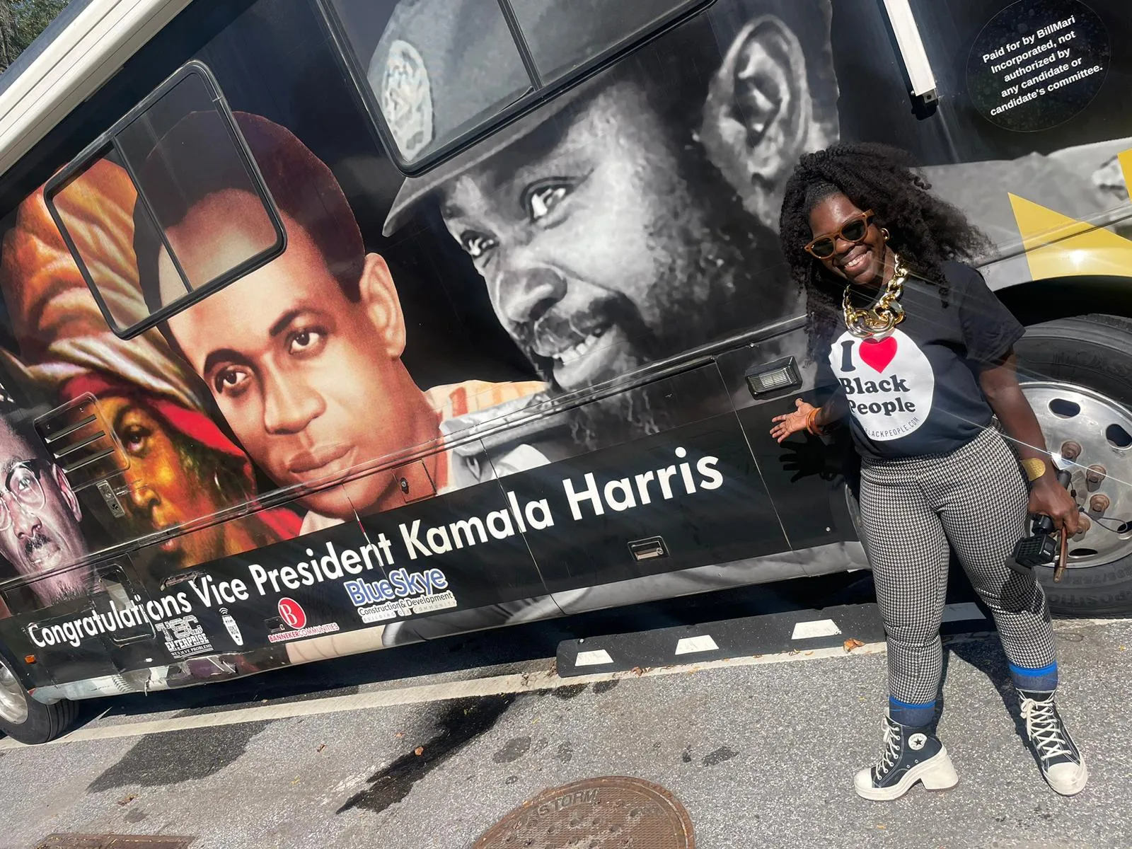 A woman wearing sunglasses, a black t-shirt saying 'I love Black People,' patterned pants, and black high-top shoes, posing and smiling in front of a bus with large portraits and the text 'Congratulations Vice President Kamala Harris.'
