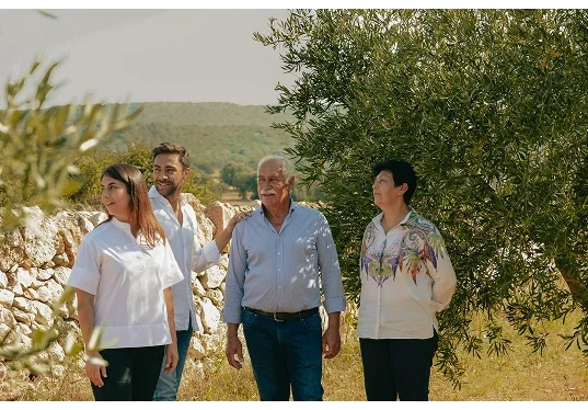 Four people standing outdoors near olive trees and a stone wall, with hills in the background.  Frisino - Masseria Vallenza