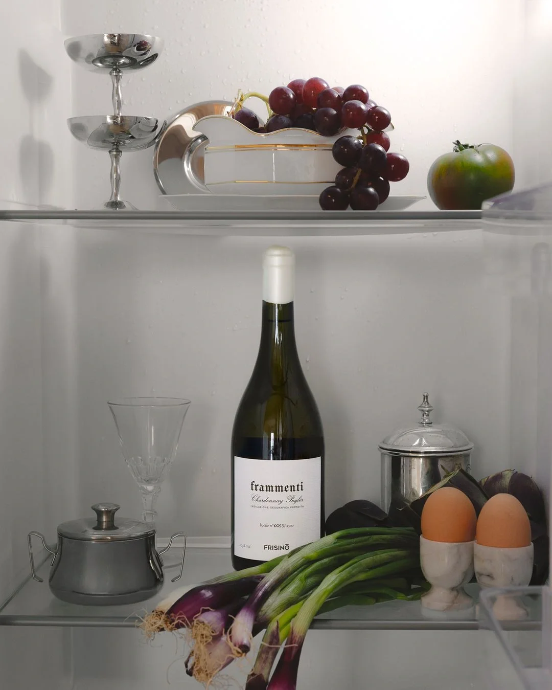 Open refrigerator shelves with a bottle of wine, two eggs in holders, green onions, purple grapes in a white dish, a green tomato, a glass, and silver containers.  Frisino - Masseria Vallenza