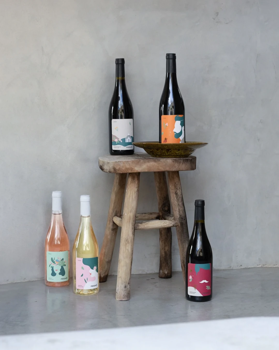 Five bottles of wine with colorful artistic labels arranged on and around a rustic wooden stool against a gray wall.  Frisino - Masseria Vallenza