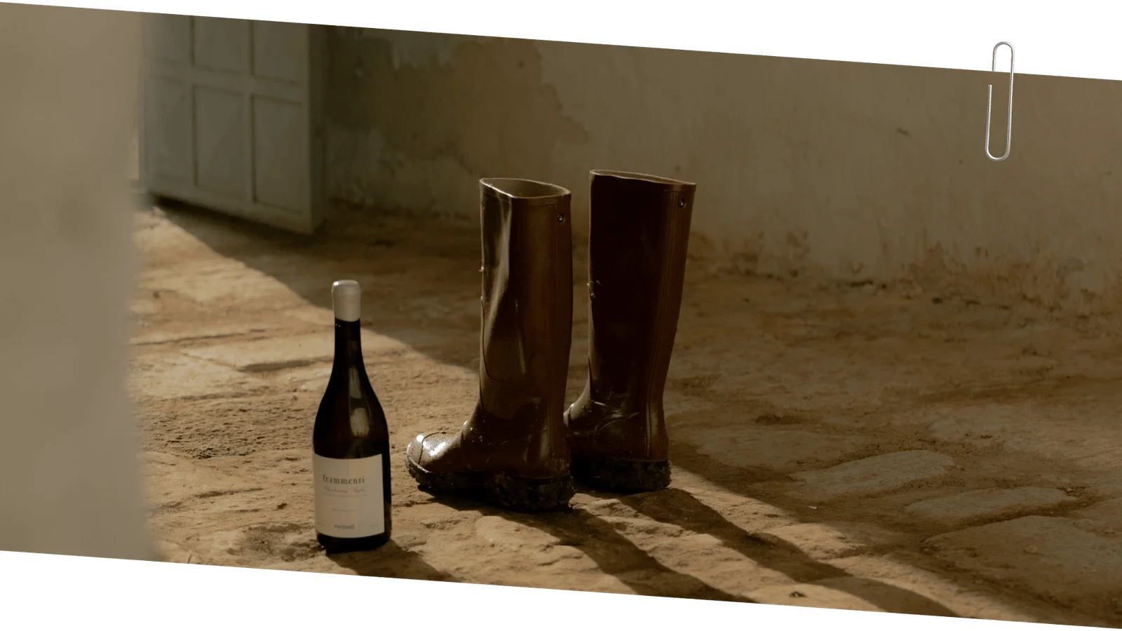 Pair of muddy brown rubber boots and a bottle of white wine on a sunlit dirt floor near an open door.  Frisino - Masseria Vallenza