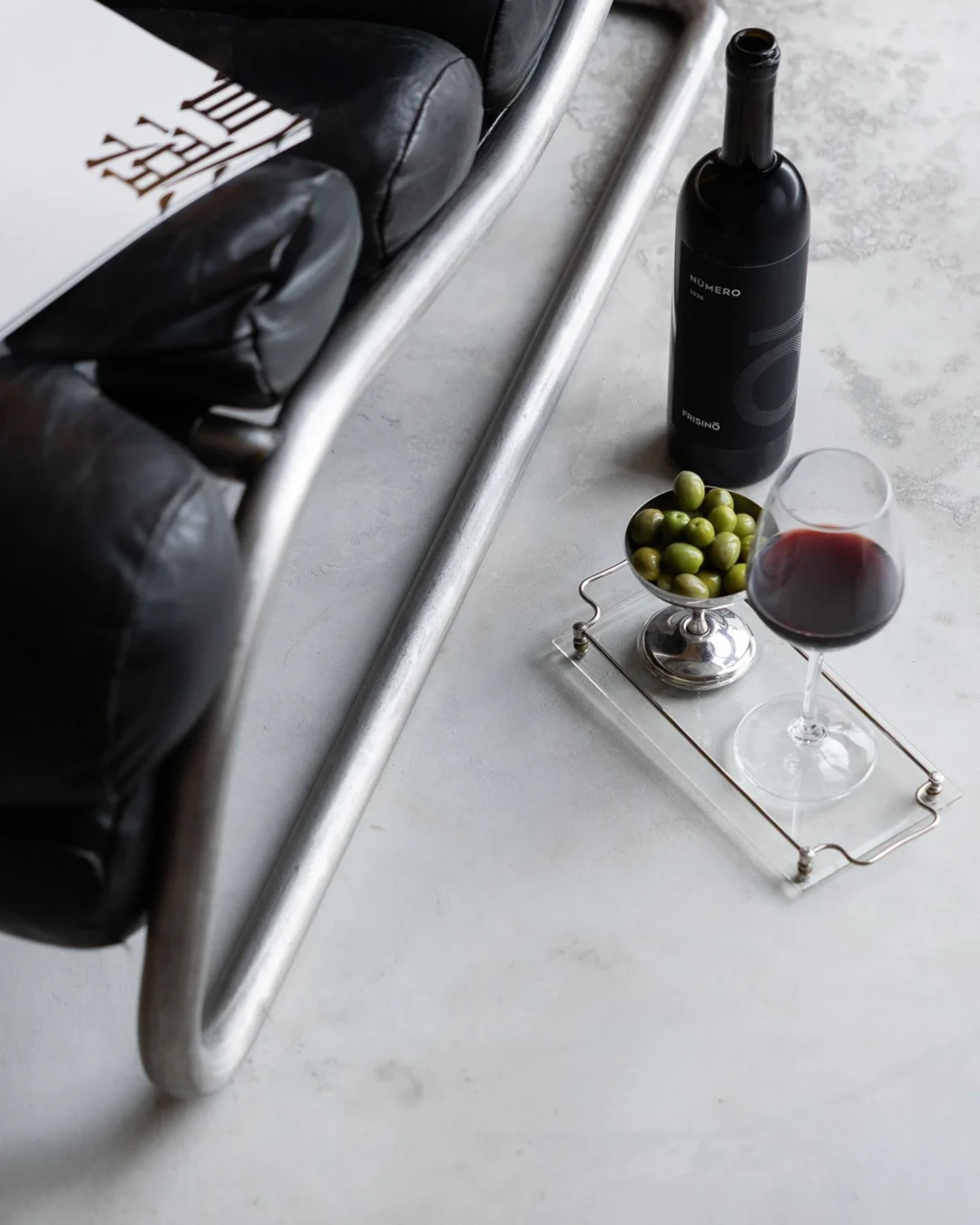 Glass of red wine, bottle of Numero wine, and bowl of green olives on a clear tray beside a black leather chair with metal frame on a gray concrete floor.  Frisino - Masseria Vallenza