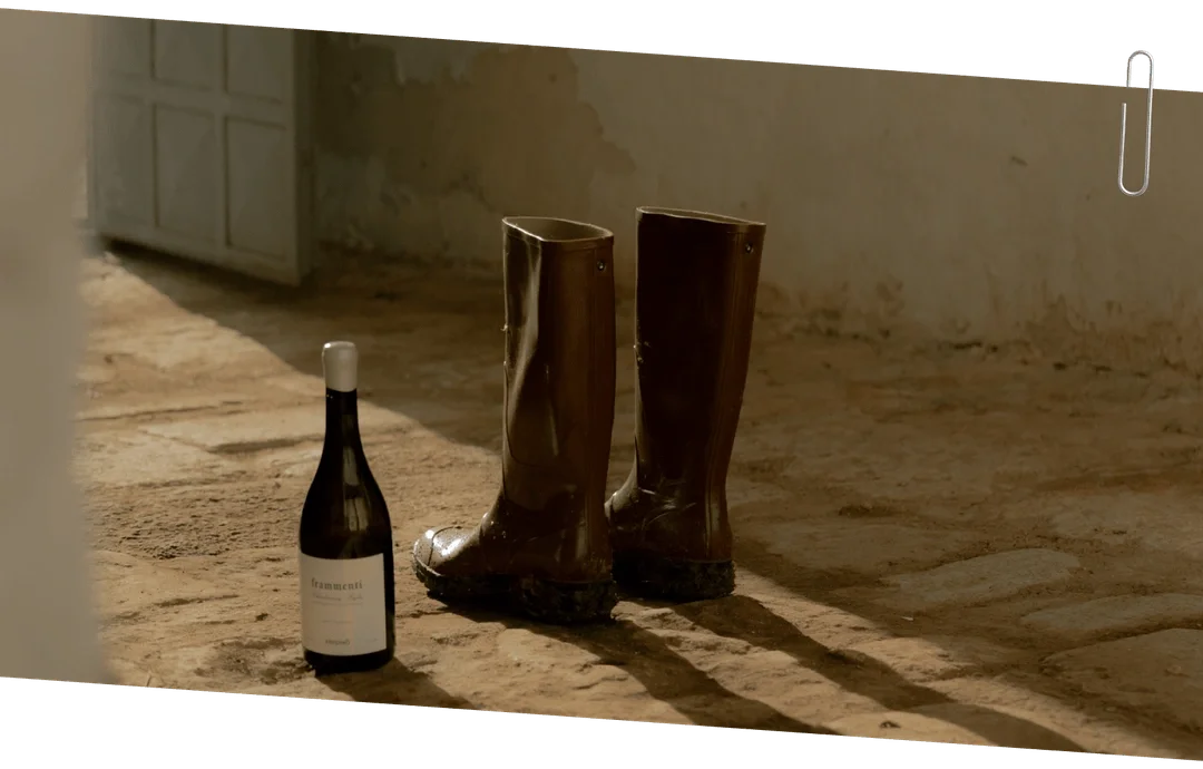 Pair of muddy brown rubber boots and a bottle of Frammenti Chardonnay white wine on a sunlit dirt floor indoors near a white door.  Frisino - Masseria Vallenza