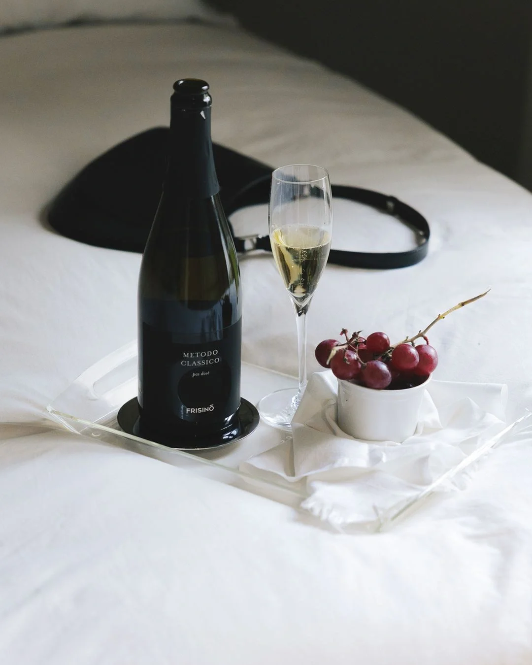 Tray on white bed with a bottle of Metodo Classico sparkling wine, filled champagne flute, and a small cup of red grapes on a white cloth.  Frisino - Masseria Vallenza