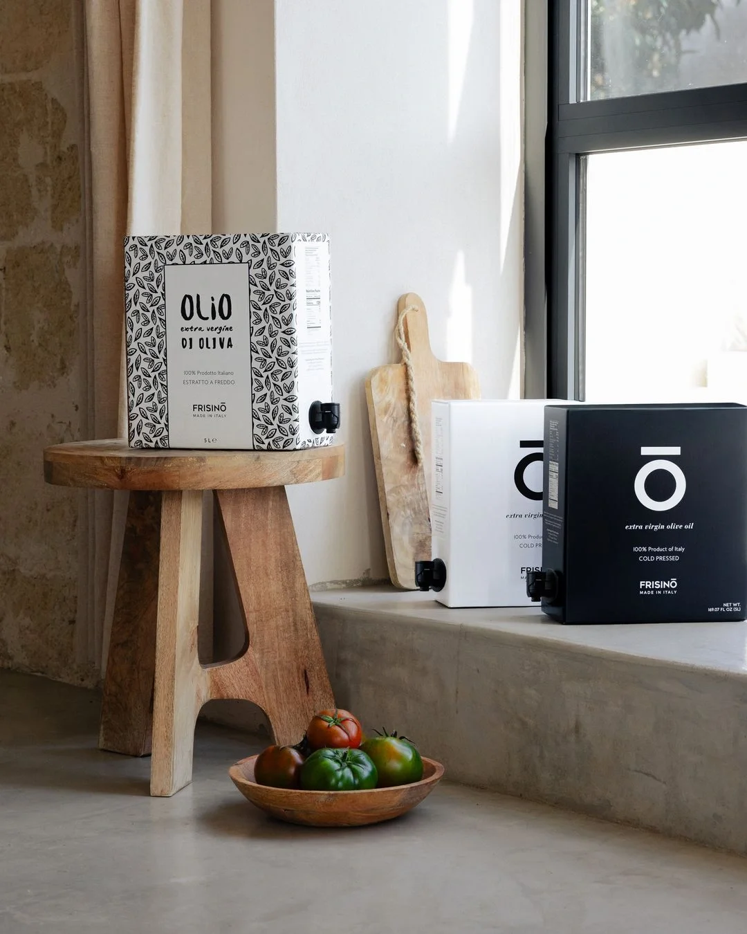 Wooden stool holding a boxed container of Italian extra virgin olive oil, next to two more boxed olive oils on a windowsill and a wooden bowl with green and red tomatoes on the floor.  Frisino - Masseria Vallenza