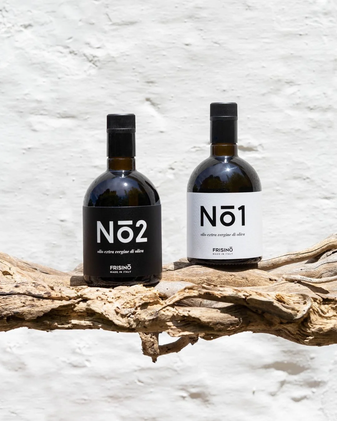 Two dark glass bottles of Frisino extra virgin olive oil labeled No1 and No2, placed on a rustic branch against a white textured wall.  Frisino - Masseria Vallenza