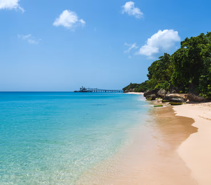 Clear turquoise ocean water gently washing onto a sandy beach with green trees on the right and a wooden pier extending into the sea under a blue sky with a few clouds.