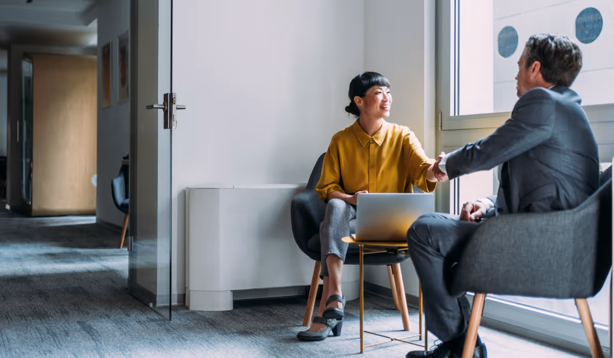 Two business professionals shaking hands while seated in an office near a window with a laptop on the table.