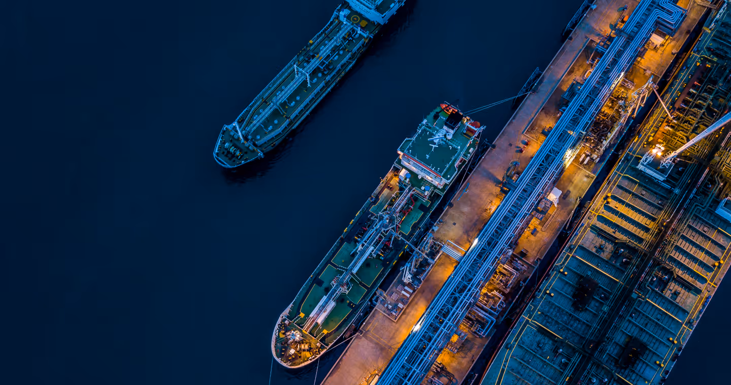 Aerial view of two large cargo ships docked at an industrial port with blue-lit pipelines and yellow lighting.