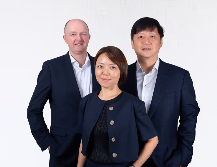 Three professionally dressed individuals standing together against a light background, two men in dark suits and a woman in a dark buttoned jacket.