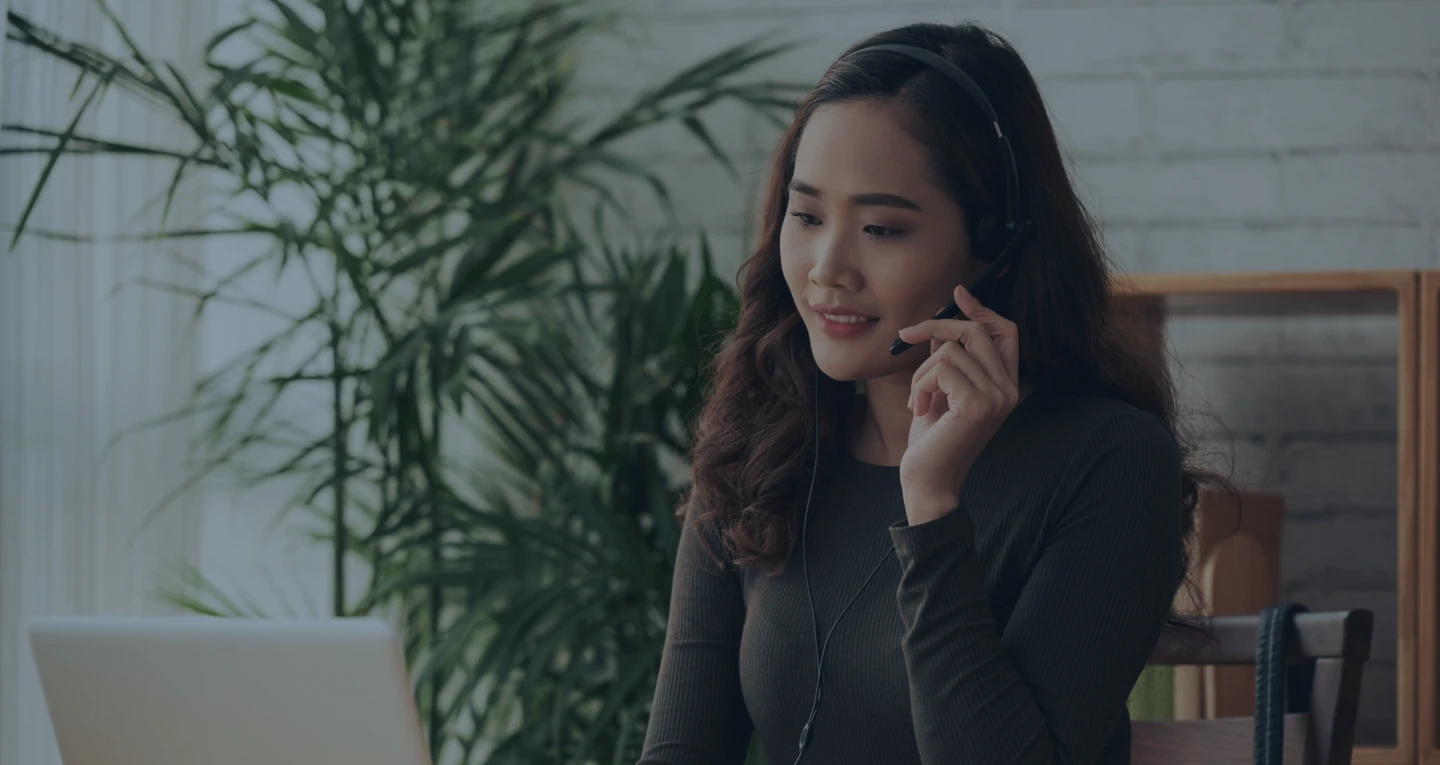 Woman with headset working on a laptop in a home office with plants in the background.