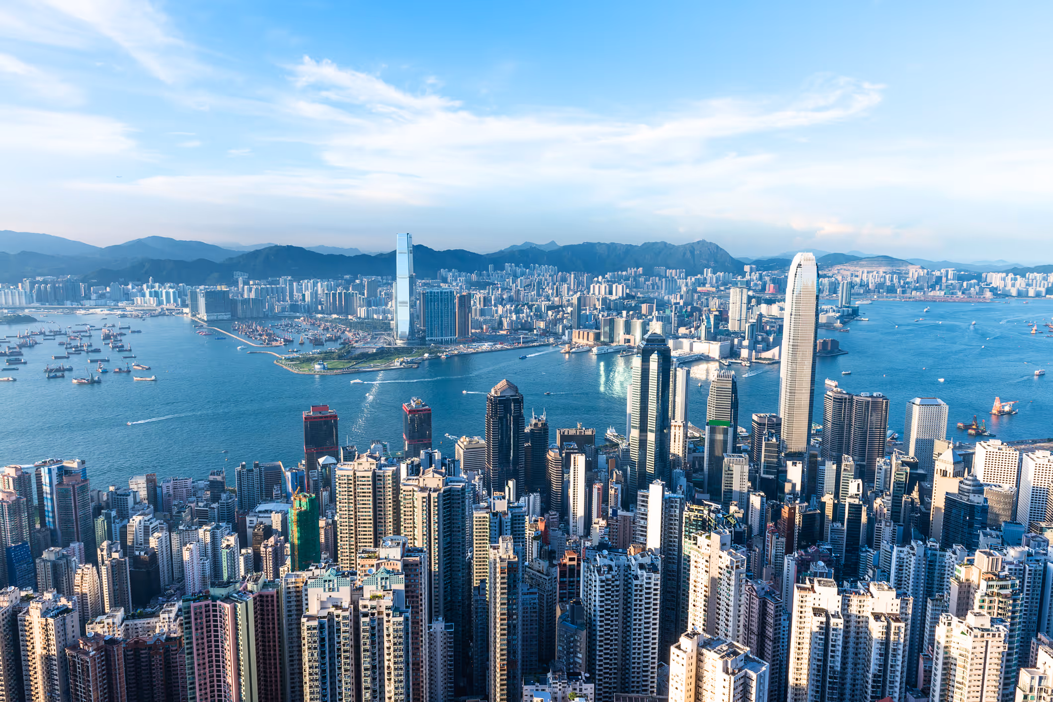 Aerial view of a dense cityscape with tall skyscrapers along a wide harbor filled with boats and mountains in the background under a partly cloudy sky.