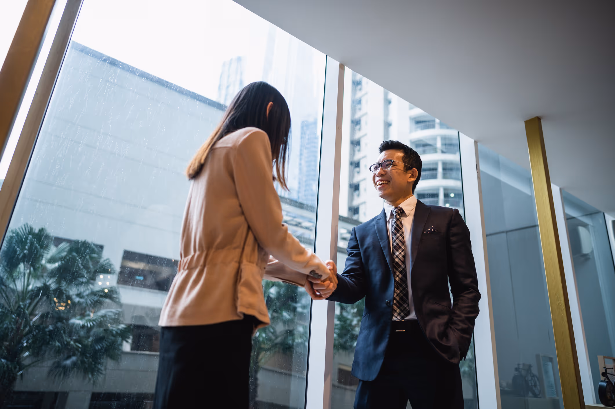 Two business professionals shaking hands near large windows in a modern office building.