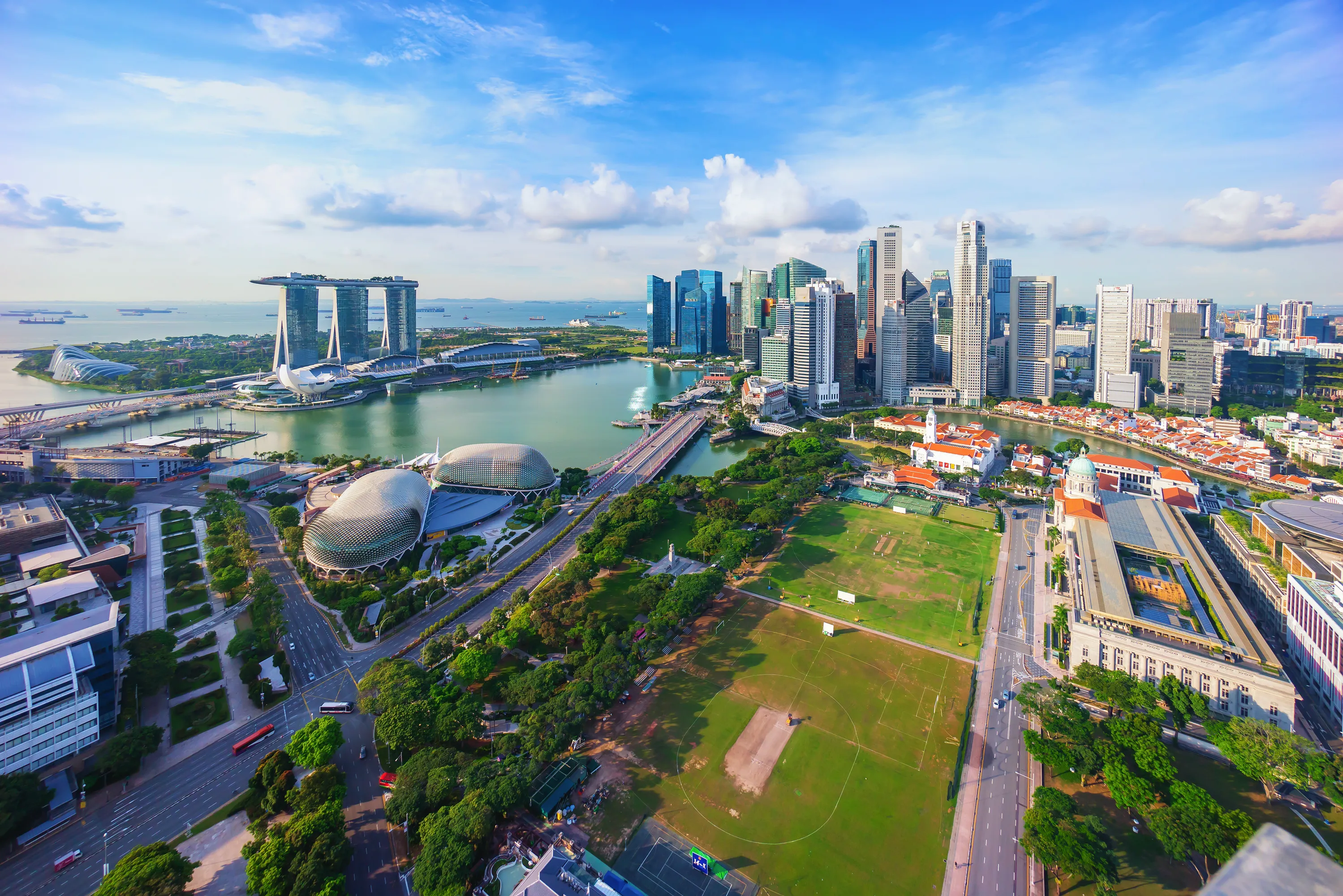 Aerial view of Singapore skyline with Marina Bay Sands, Esplanade theaters, green parkland, and downtown skyscrapers under a blue sky with scattered clouds.
