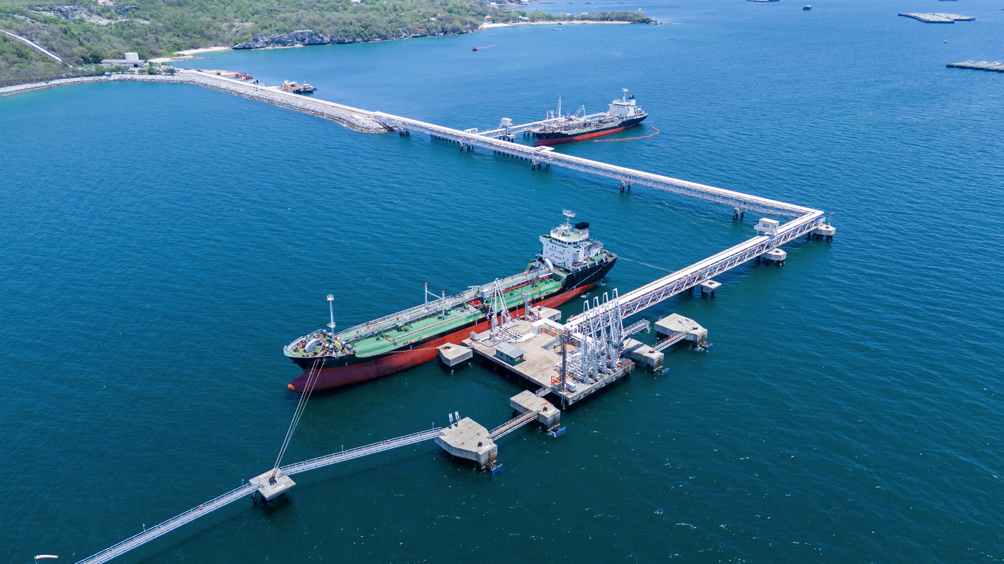 Aerial view of two large cargo ships docked at a marine oil terminal with connected piers extending from a green forested coastline.
