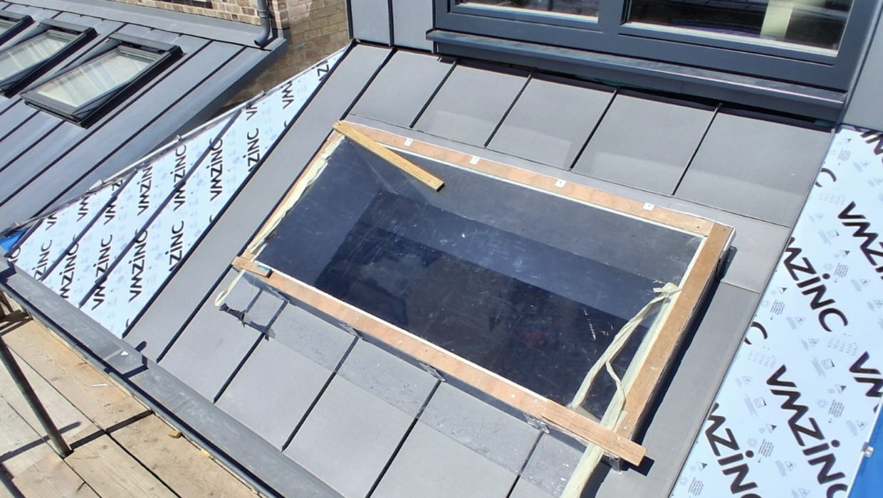 Close-up of a grey tiled roof with a rectangular skylight window framed by wood, some roofing insulation material labeled VMZINC is visible around the window.