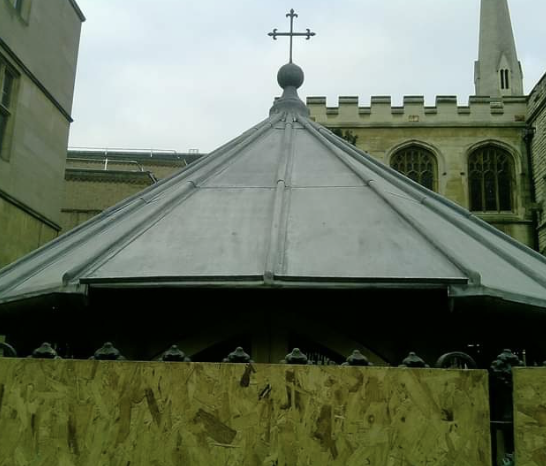 Steeple with a cross on top behind a wooden fence and historic stone building with arched windows.