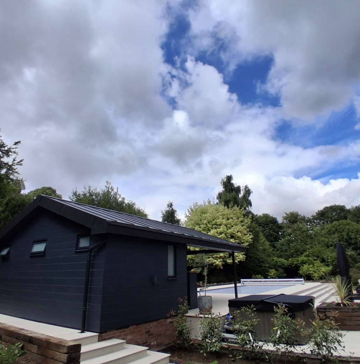 Modern dark blue pool house next to a covered hot tub and an empty outdoor swimming pool surrounded by trees under a partly cloudy sky.