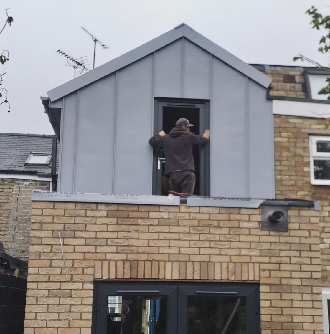 Man installing or adjusting a door in a gray dormer extension above a brick building.
