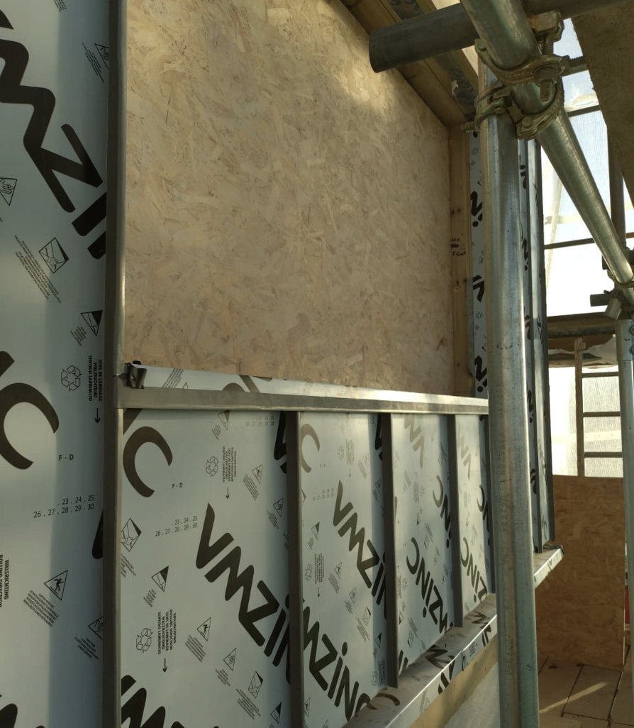 Partially constructed wall with OSB sheathing and protective membrane branded VMZINC, surrounded by metal scaffolding.