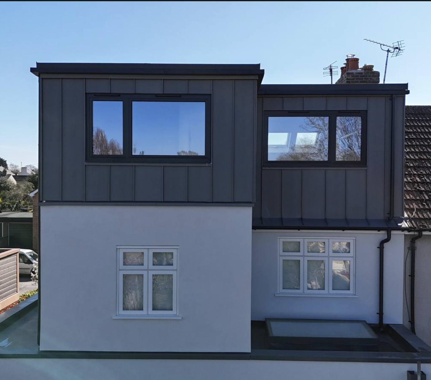 Modern two-story house extension with dark cladding on the upper floor and white walls below, featuring multiple windows reflecting a clear blue sky.