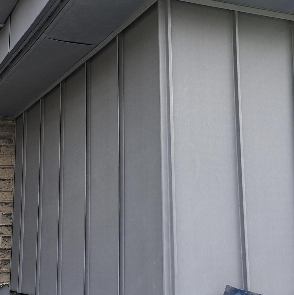 Gray metal siding panels on a building corner with a brick wall partially visible on the left.