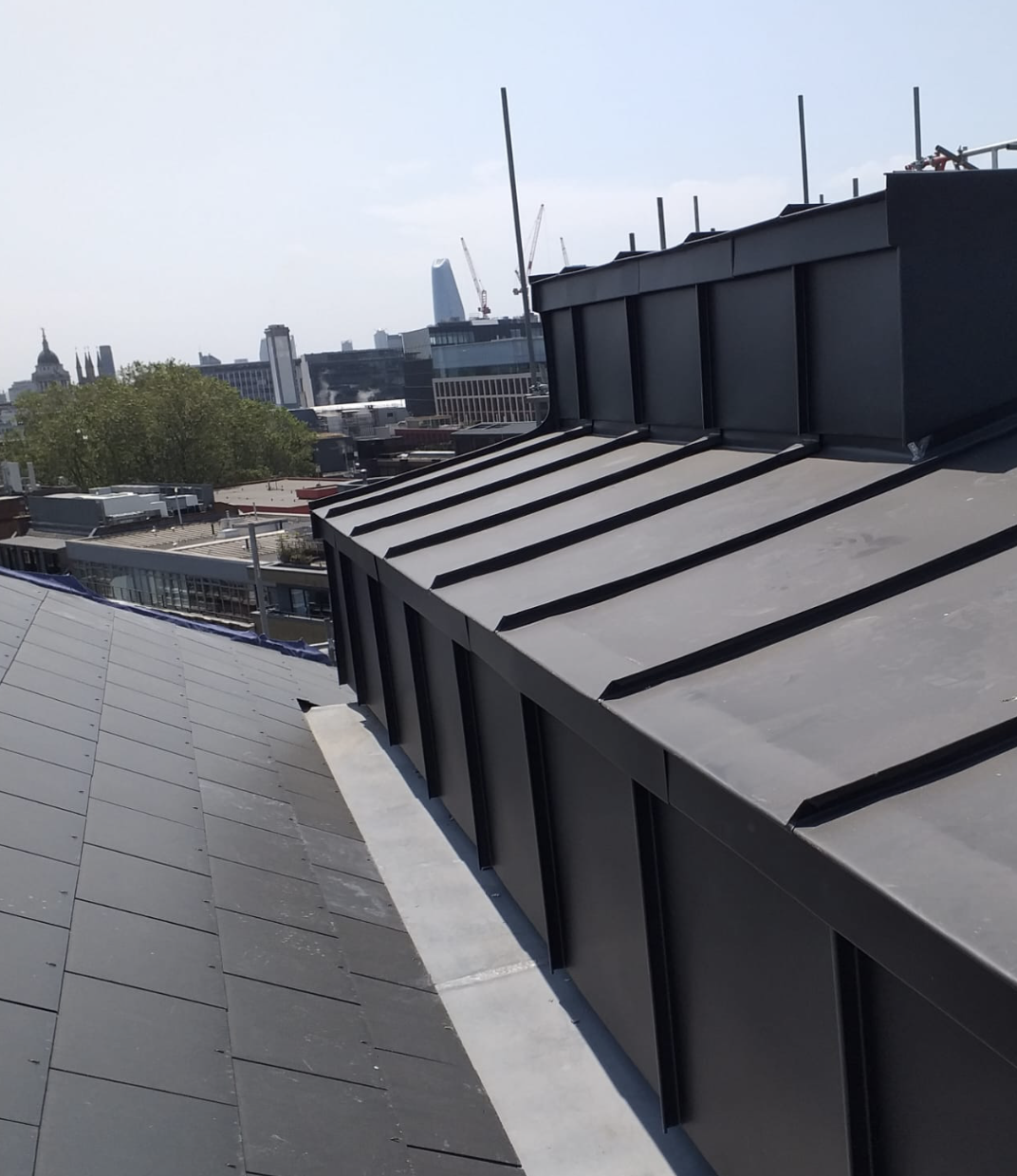 View of a sloped rooftop with dark metal panels and urban buildings with cranes in the background under a clear sky.