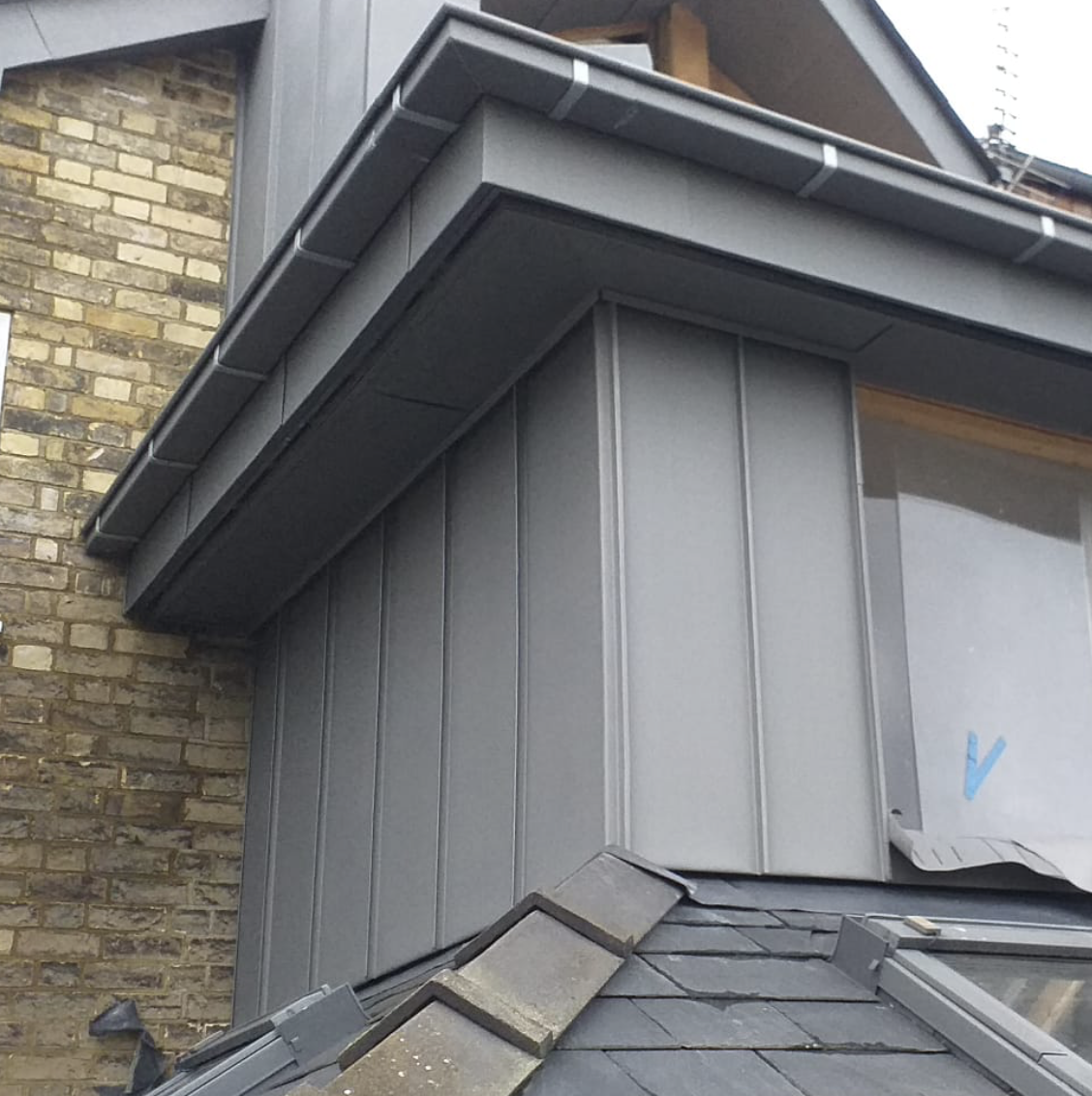Close-up of a building's corner showing grey metal cladding, roof edge gutter, and brick wall background.