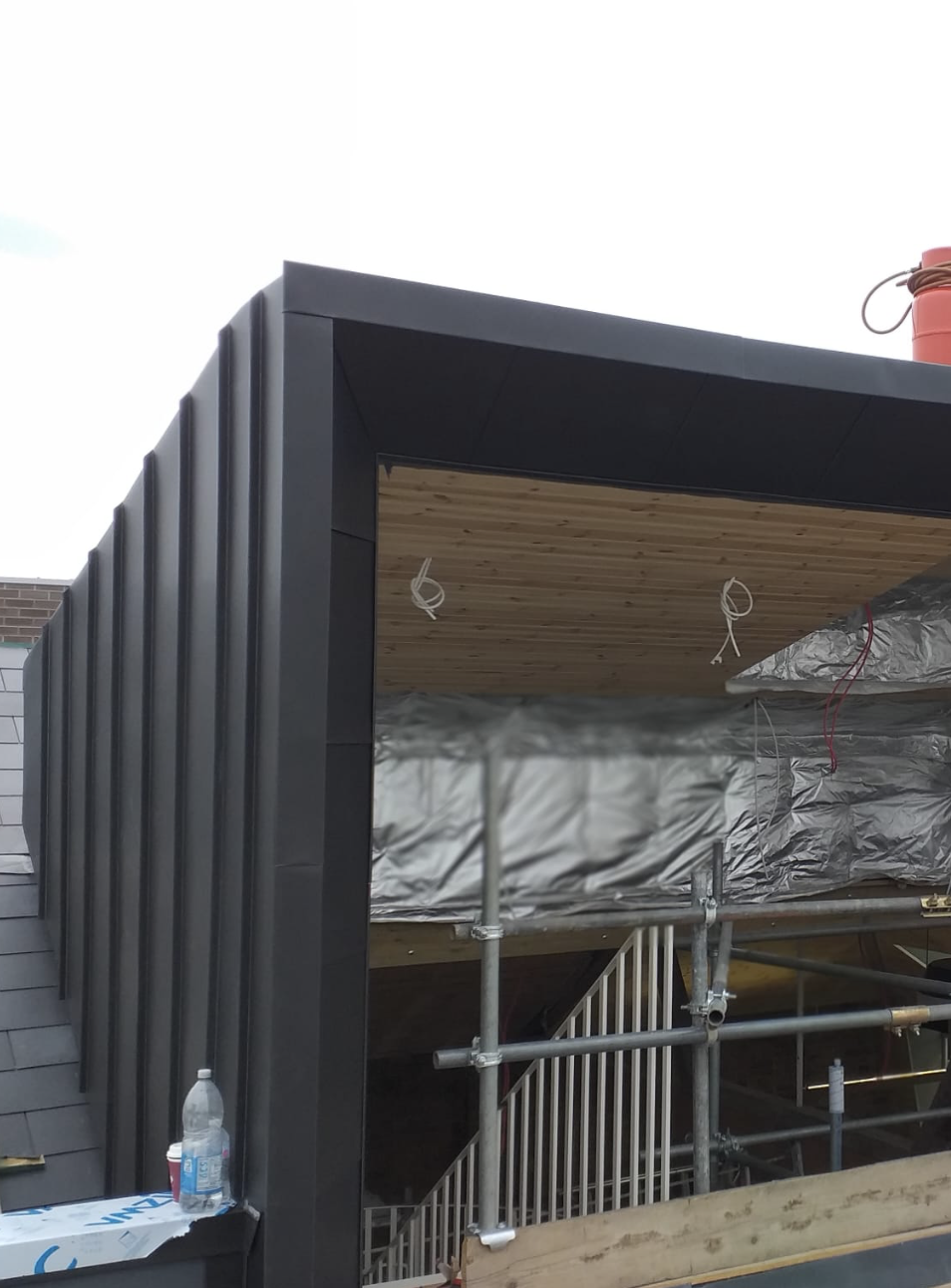 Construction site showing a modern building exterior with black metal siding, wooden ceiling, scaffolding, and insulation materials inside.