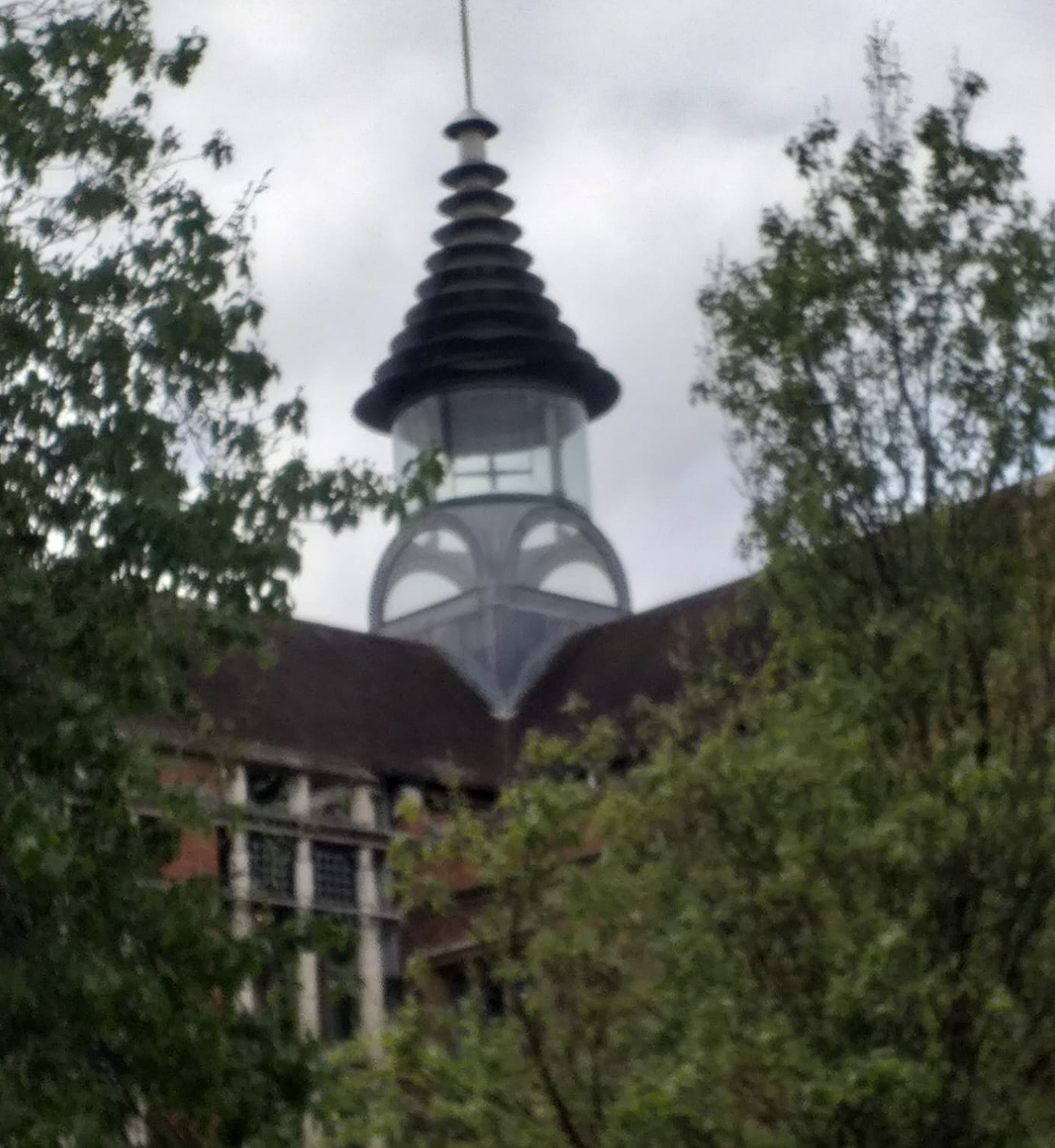 Tapered tower with layered black roof and arched windows, partially obscured by green leafy trees.