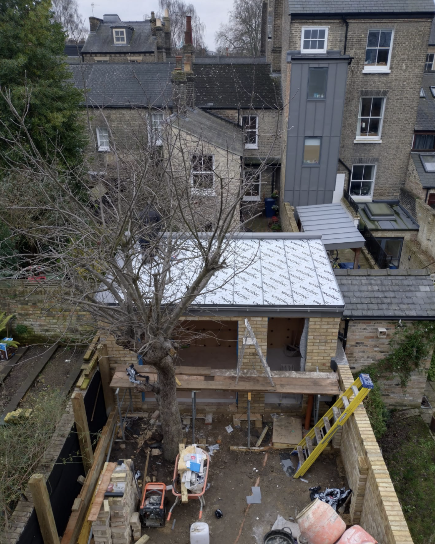 Construction site in a backyard with a newly built brick structure, a bare tree in front, ladders, and building materials scattered around.