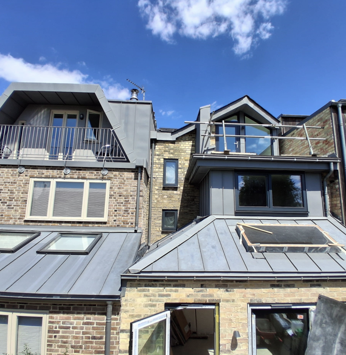 Modern brick townhouse with angled grey metal roofs, balconies, large windows, and an open door under a blue sky with scattered clouds.