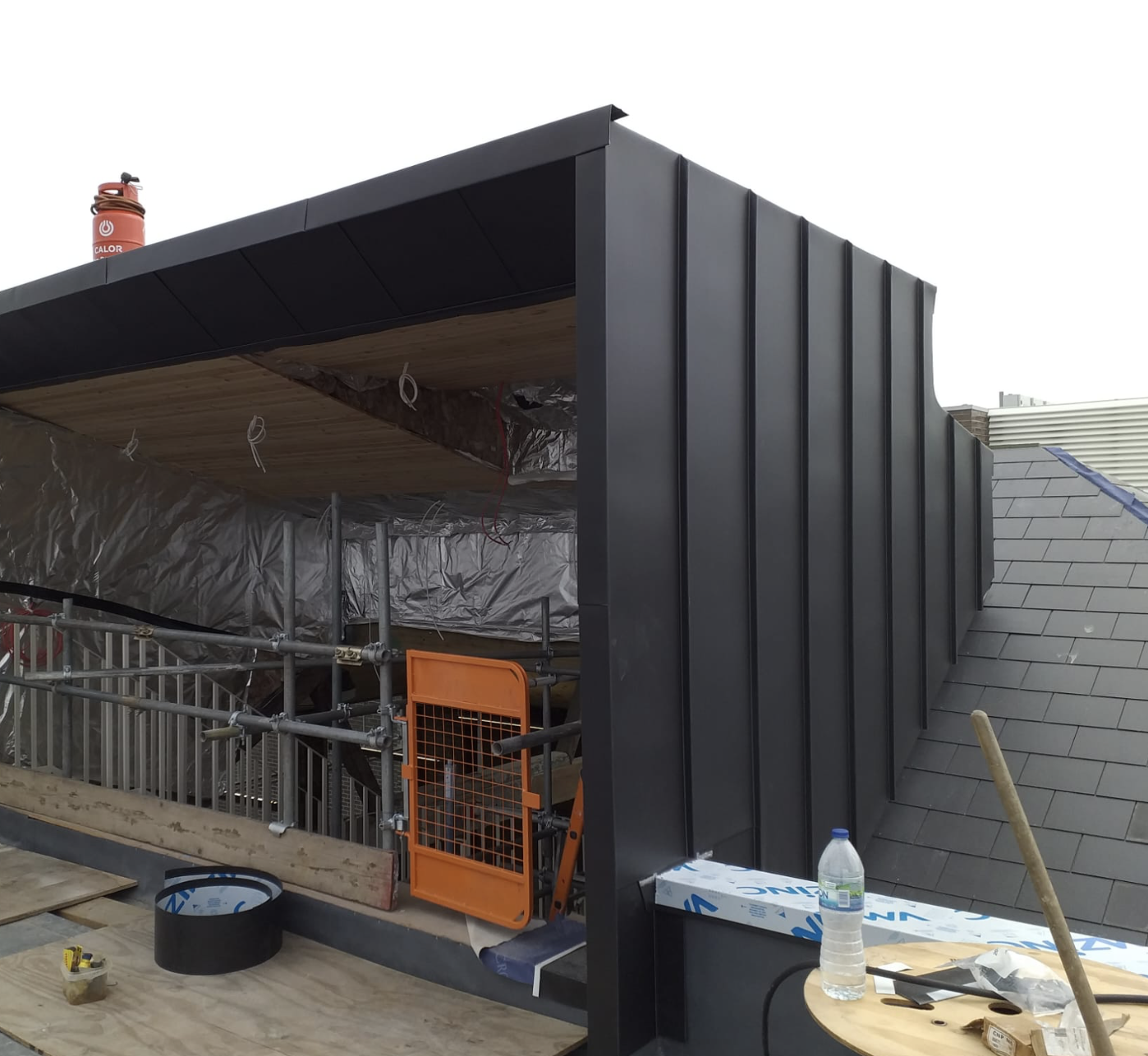 Partially enclosed rooftop structure under construction with black metal cladding, scaffolding, orange safety gate, and construction materials.