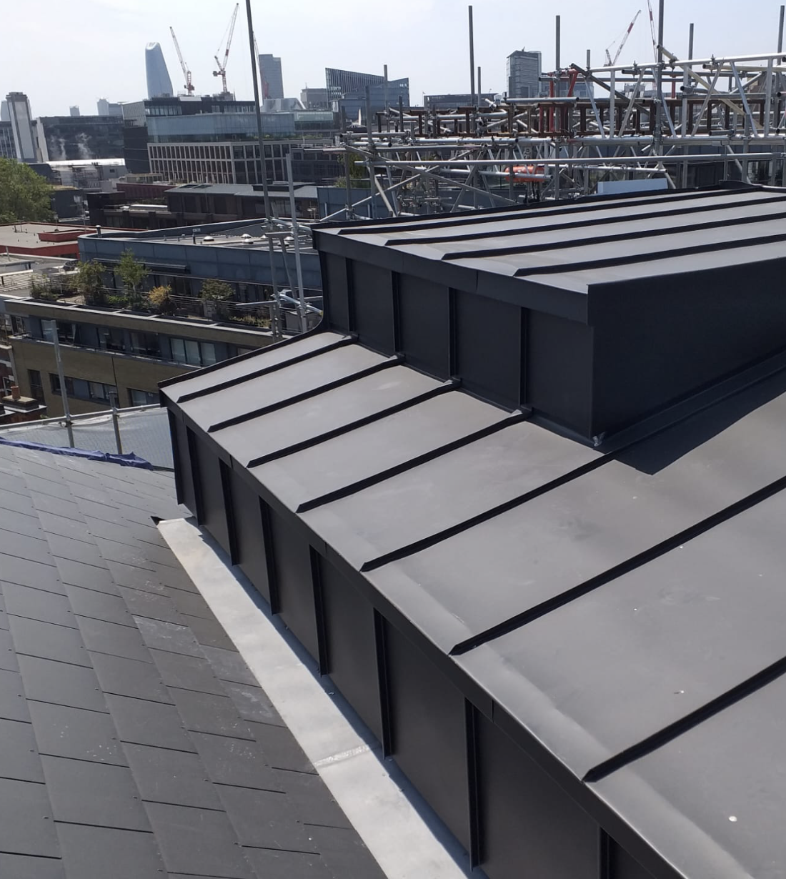 Close-up of a modern dark metal rooftop with structural beams, with a city skyline and construction cranes in the background.