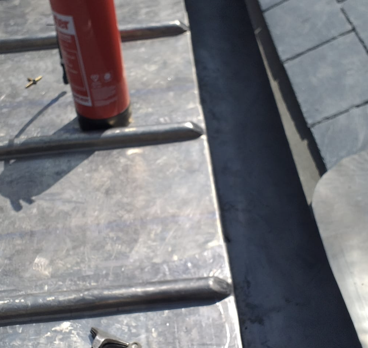 Red fire extinguisher standing on a metallic surface with ribbed bars and a screwdriver.