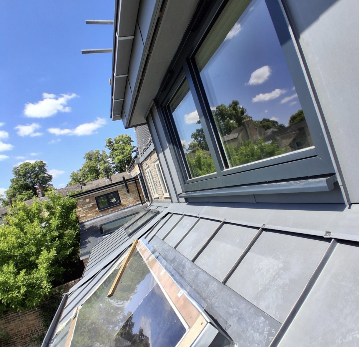 Close-up of modern grey metal roof with skylight and windows reflecting blue sky and trees.