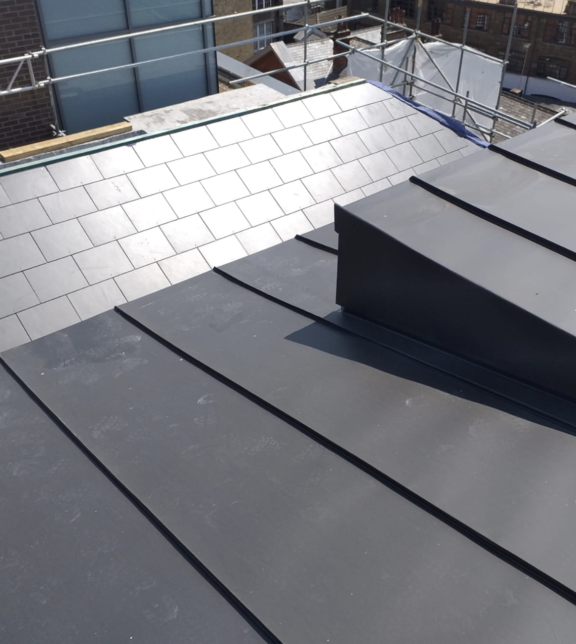 Roof with dark metal standing seam panels and a section of light gray roof tiles under construction with scaffolding in the background.