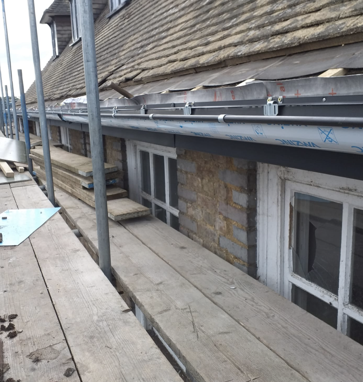 Wooden scaffolding along the side of a building with a sloped roof under repair and a broken window below.