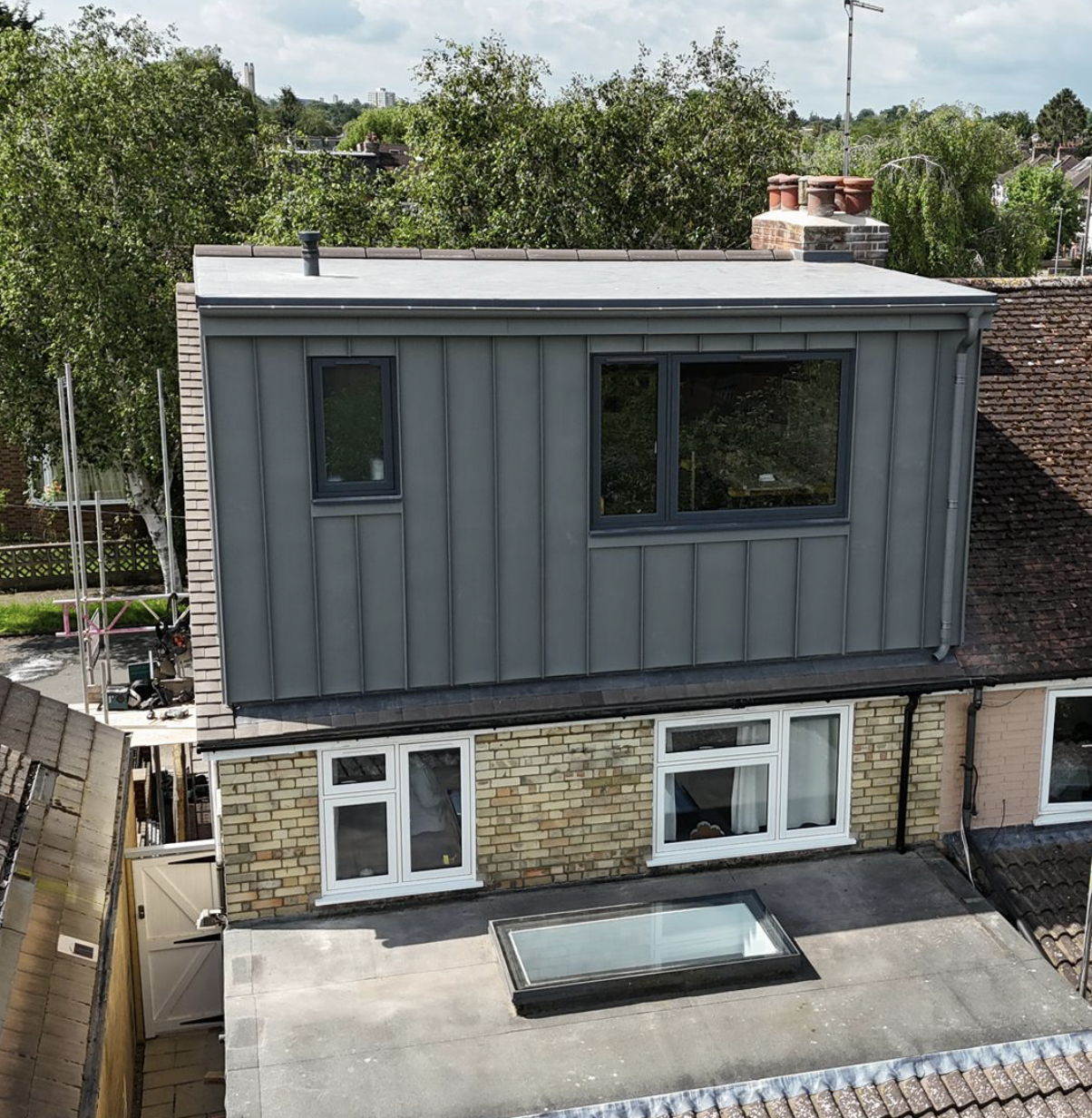 A modern gray extension with large windows added above a brick house with a flat roof and skylight.